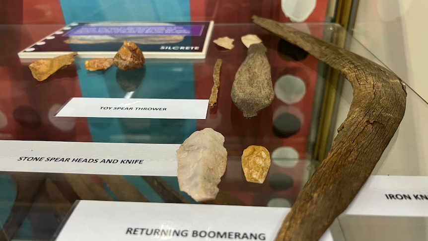 A series of ancient indigenous tools, including a toy spear launcher, in a glass cabinet. 