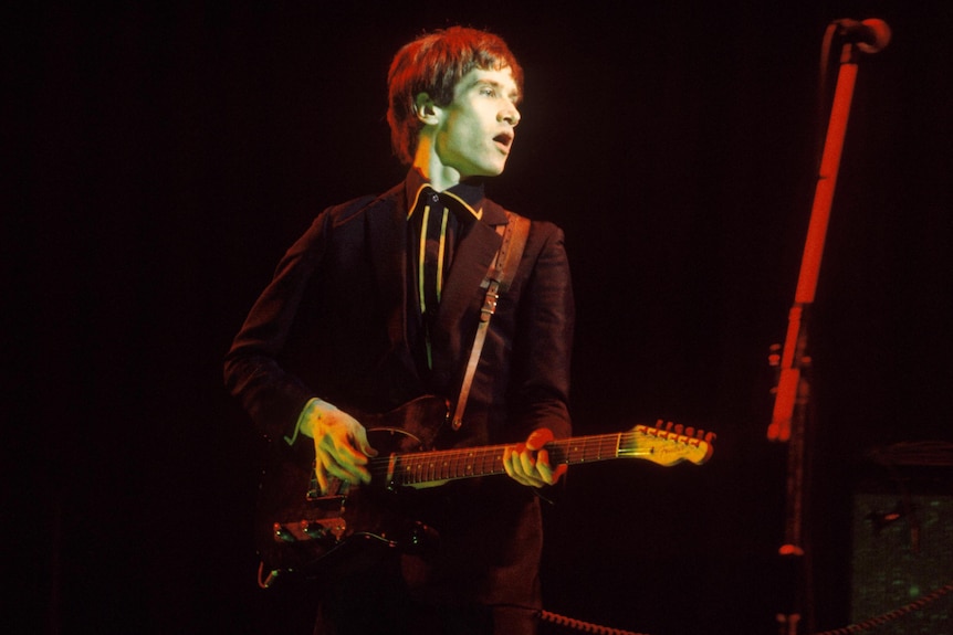 A young Wilko Johnson in dark suit playing guitar on stage with Dr. Feelgood
