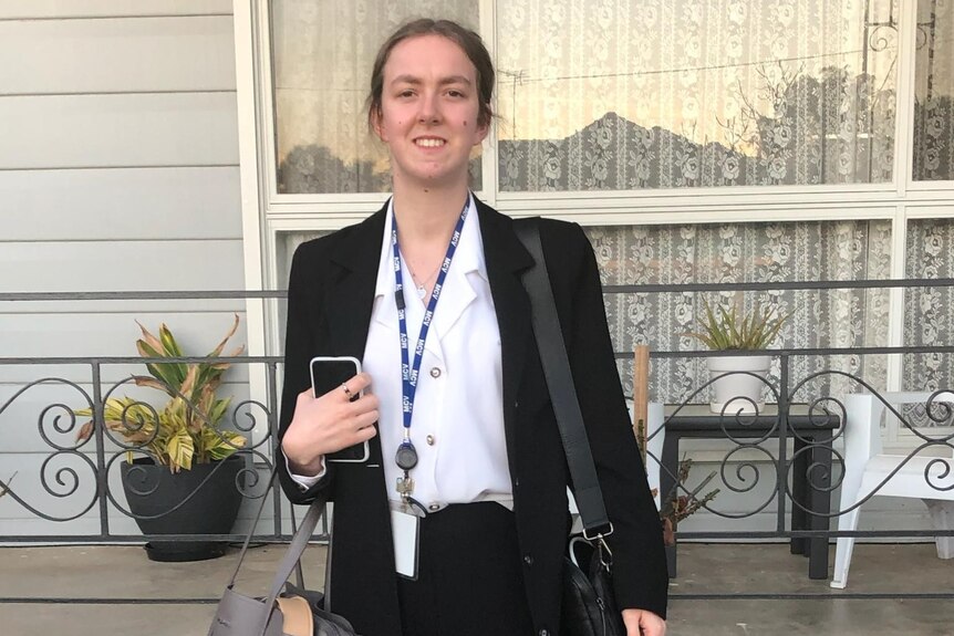 A young, brown-haired woman in a black suit and white shirt with a lanyard over it stands in front of a house