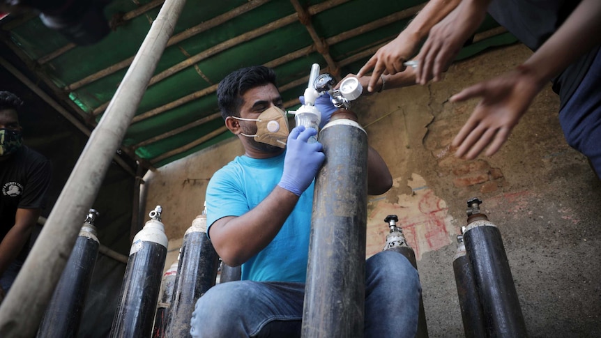 A man wearing a mask and dressed in a blue shirt and jeans holds an oxygen tanks while surrounded by other oxygen tanks.