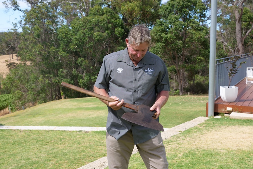 A man with grey hair, wearing a grey shirt, with a logo, stands in a garden and looks at an axe.