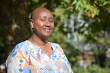 Woman smiling at camera, she has shaved head, wears colourful v neck cotton top and gold hoop earrings.