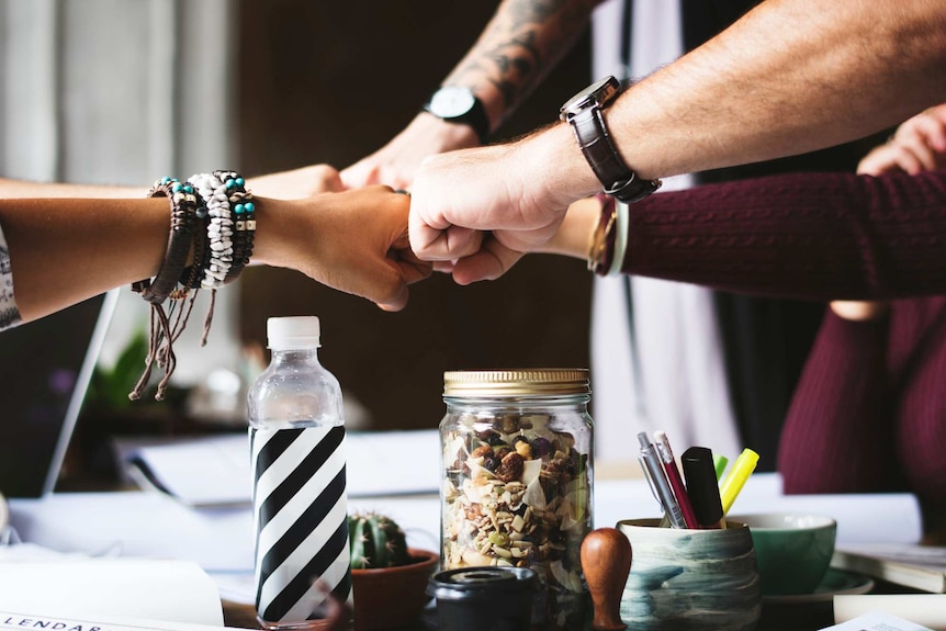 Five people pump their fists together in an office setting, depicting good and healthy work environment.