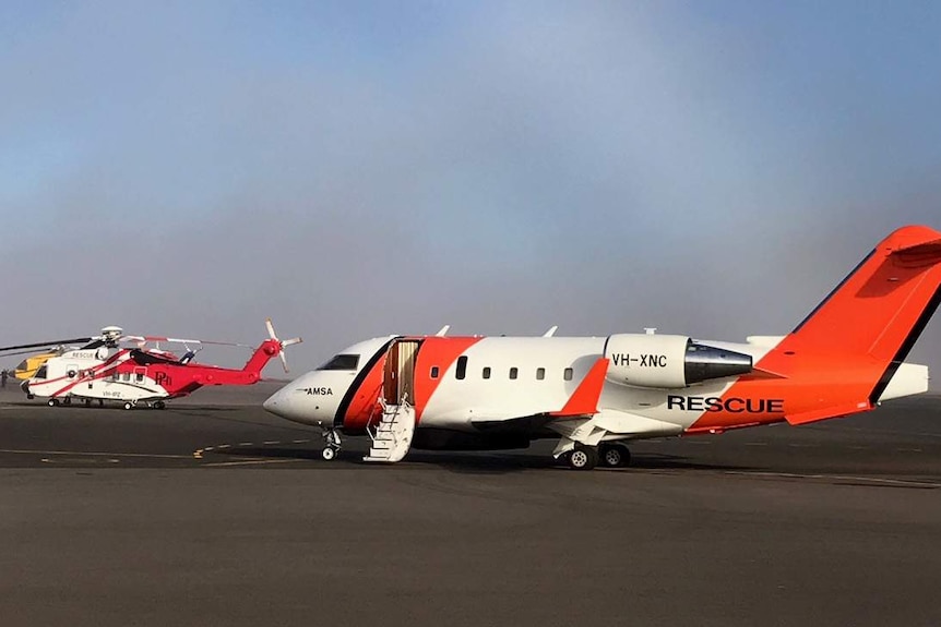 An AMSA jet and a rescue helicopter sit on the tarmac under grey skies.