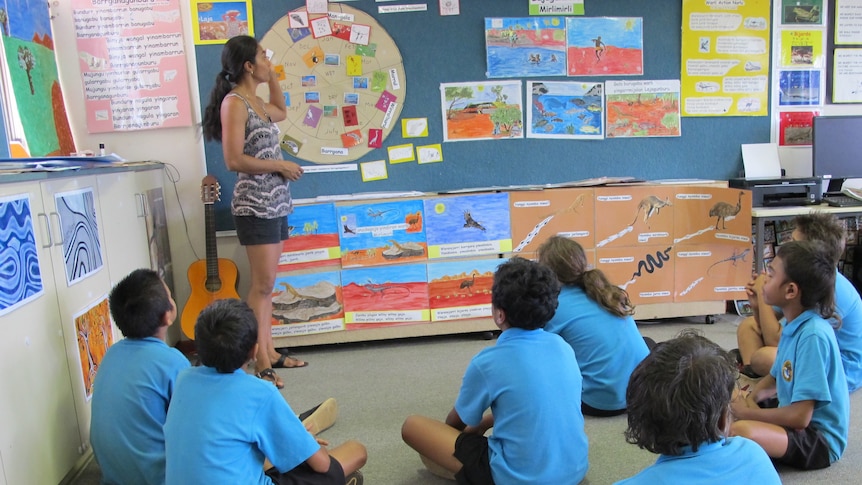 Students sit on the floor while a teacher points to colourful pictures