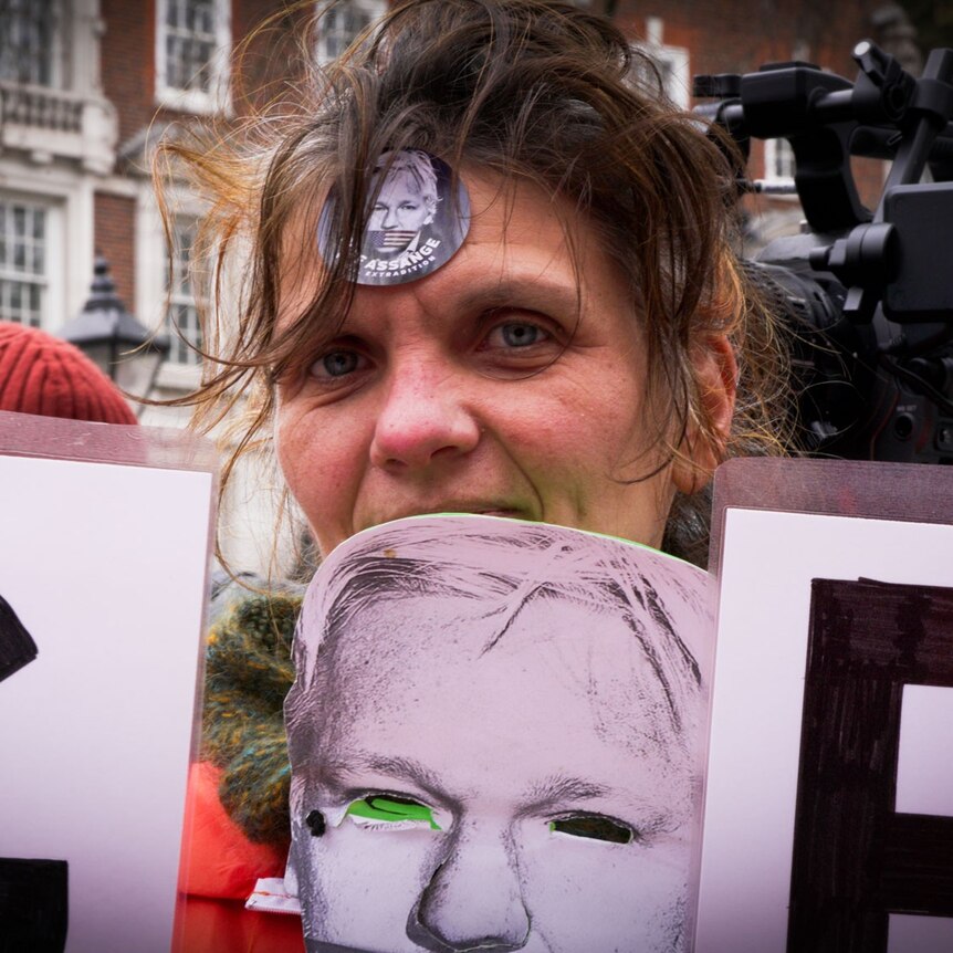 A woman with a sticker on her forehead and mask showing a man's face strung around her neck looks at the camera