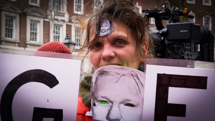 A woman with a sticker on her forehead and mask showing a man's face strung around her neck looks at the camera