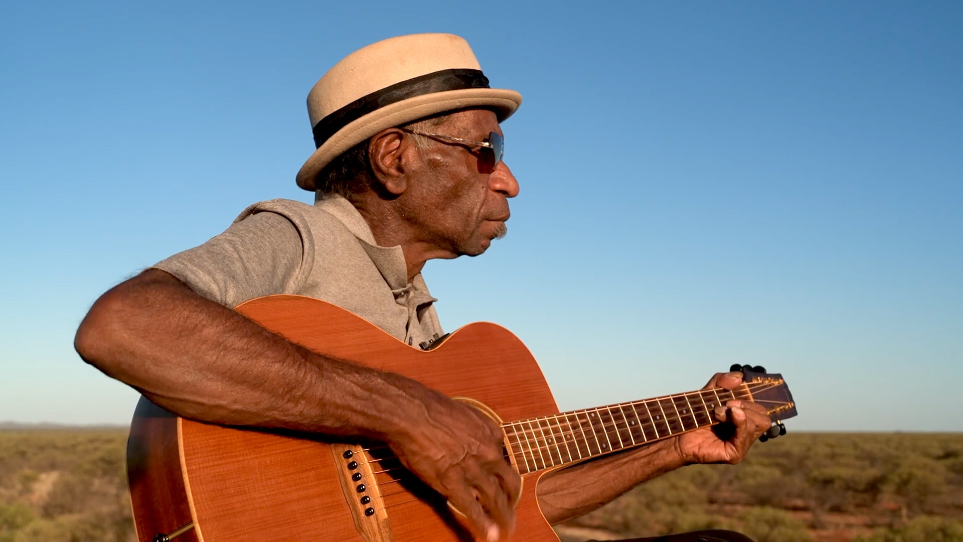 Indigenous man sits outdoors wearing a white hat and holding a guitar.
