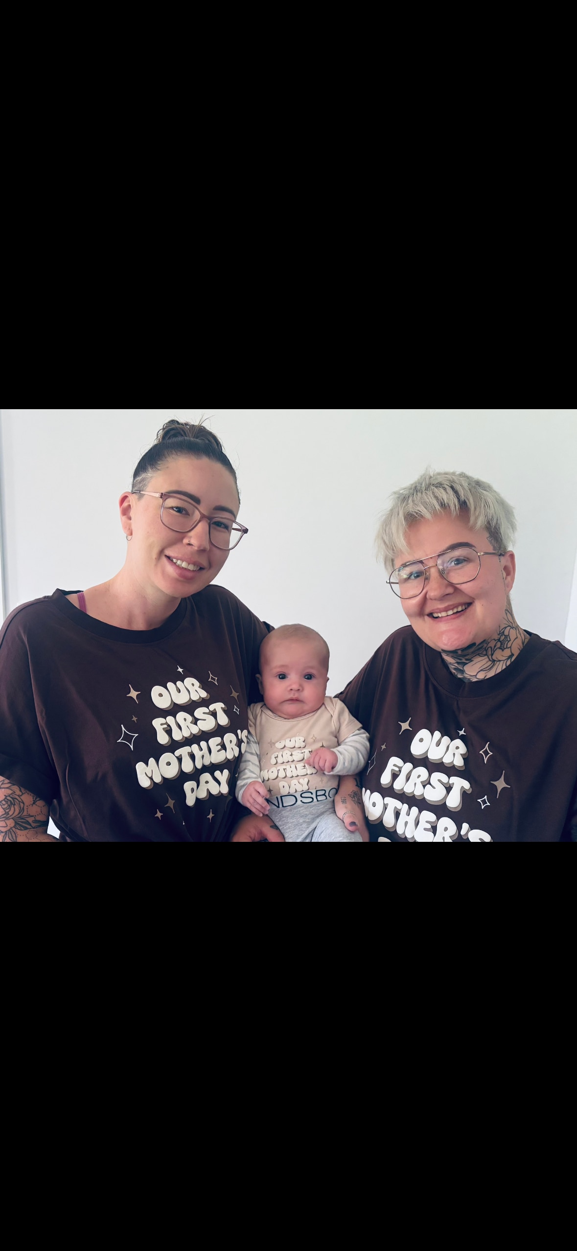 Two women wearing black Mother's Day T-shirts holding a baby between them.
