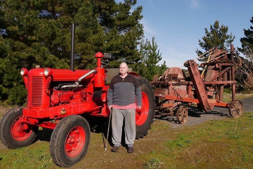 An elderly man stands in front of a bright red tractor with a wooden chaff cutter attached.