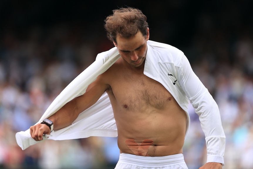 A male tennis player takes off his white shirt to show his bare torso
