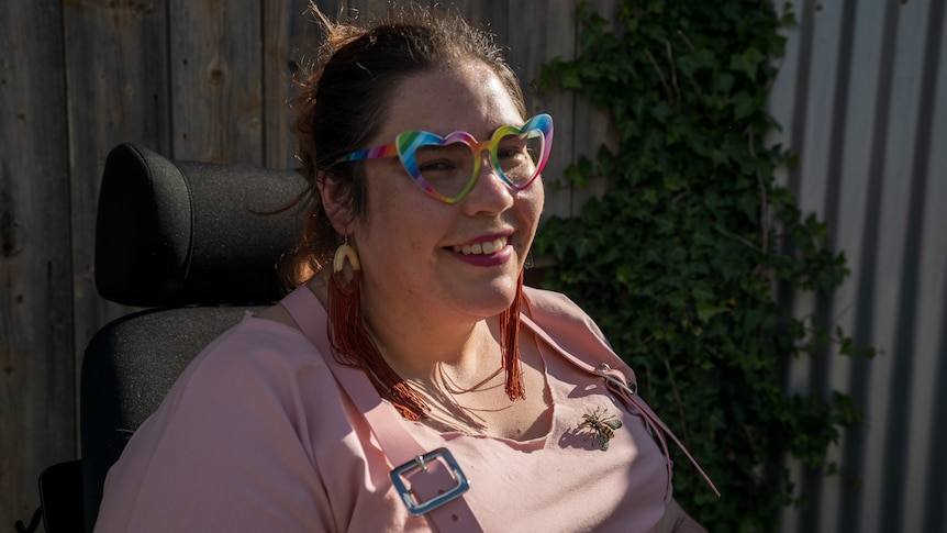 A woman with a pink shirt and heart-shaped rainbow-colored glasses
in a wheelchair.