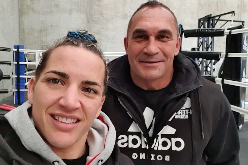 A picture of a man and woman in exercise clothing in front of a boxing ring, with exercise equipment behind them.