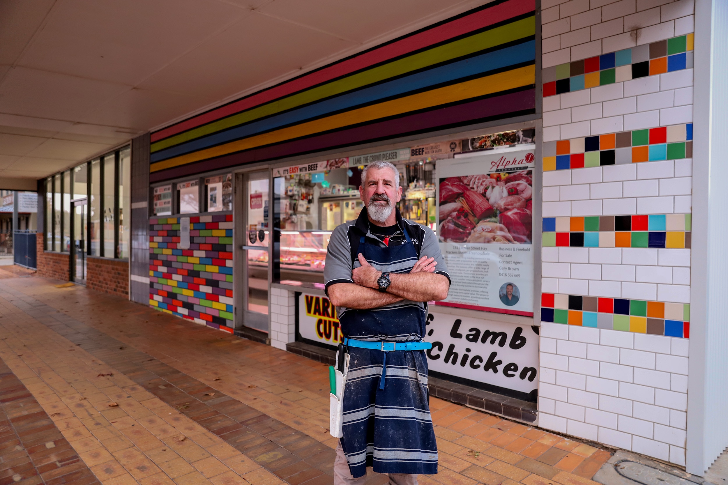 A man wearing a blue and white striped apron stands with his arms crossed outside a butcher shop with signs for meat behind him.