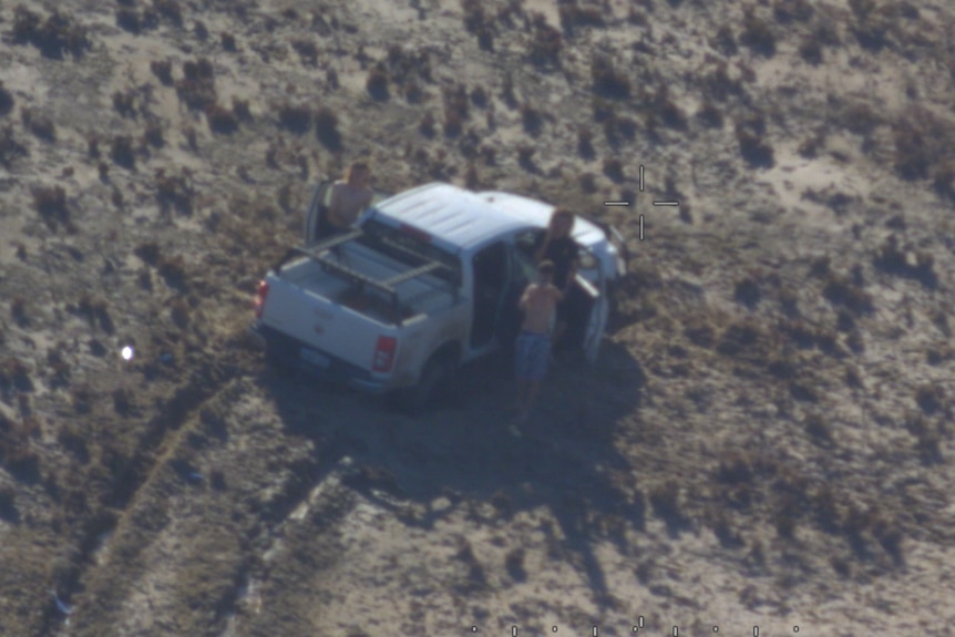 An aerial photograph of a bogged car with three teenagers standing around.  