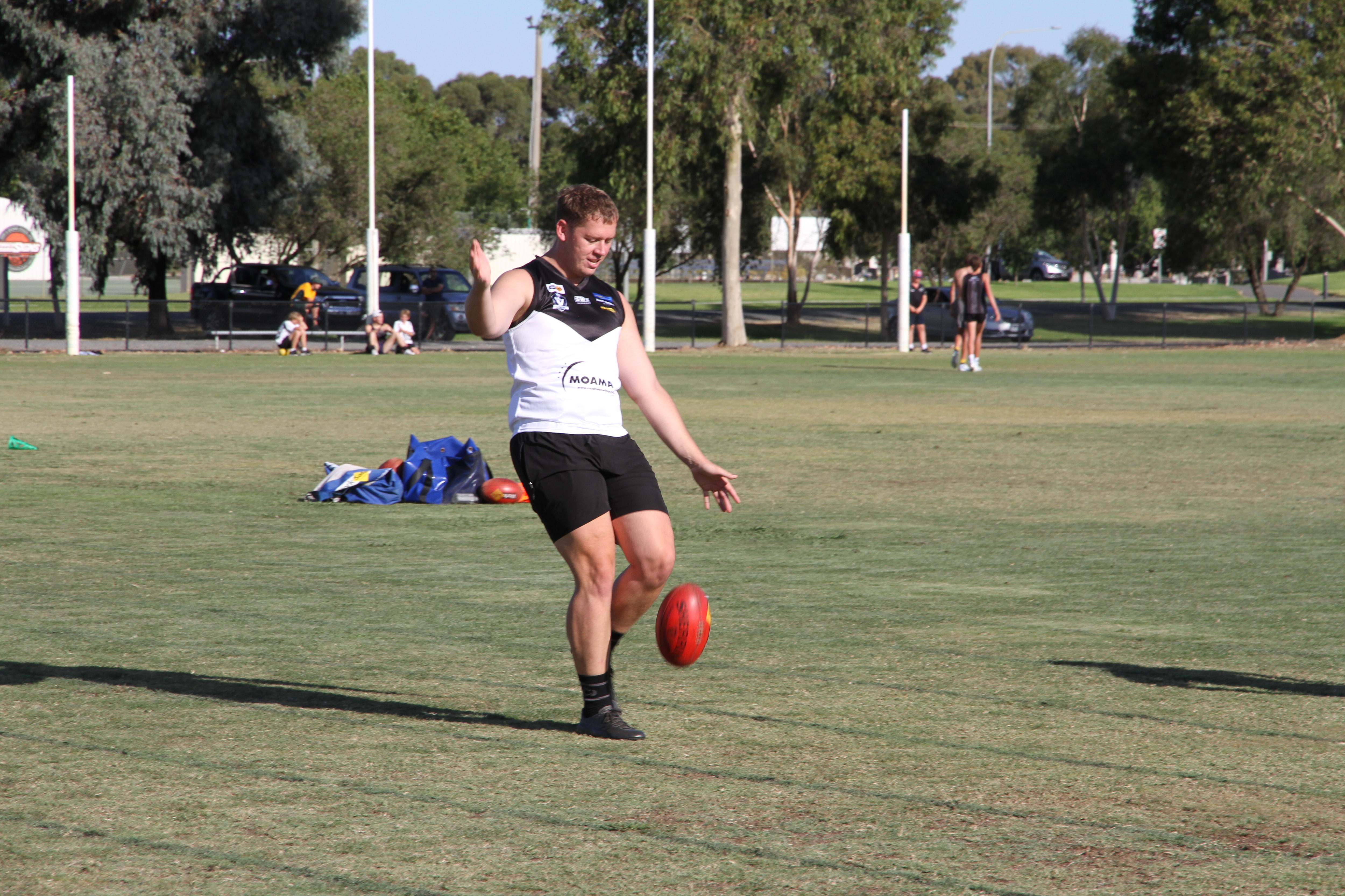 A man kicks an Australian Rules football, with others training in the background.