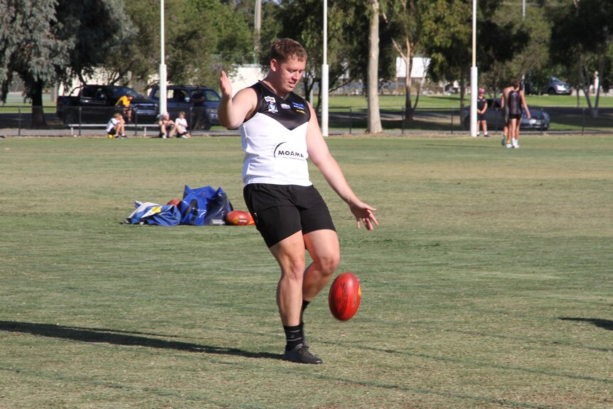 A man kicks an Australian Rules football, with others training in the background.