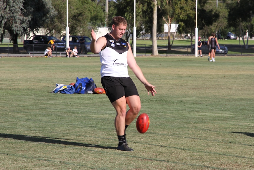 A man kicks an Australian Rules football, with others training in the background.