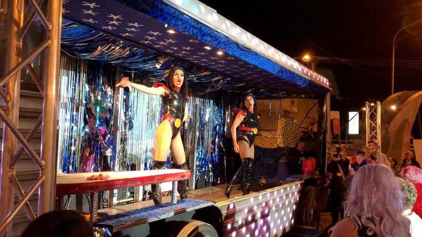 Two Indigenous drag queens perform in leotards featuring the Indigenous flag on a stage decorated with silver tinsel.