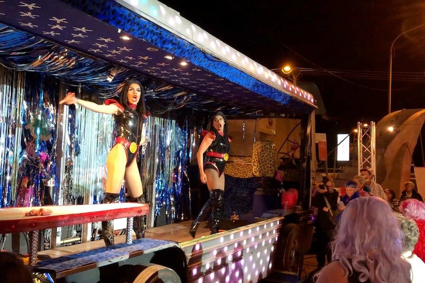 Two Indigenous drag queens perform in leotards featuring the Indigenous flag on a stage decorated with silver tinsel.