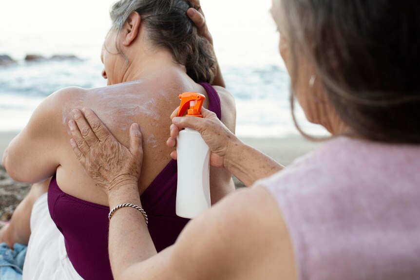 A lady applying sunscreen to another lady's back at the beach.