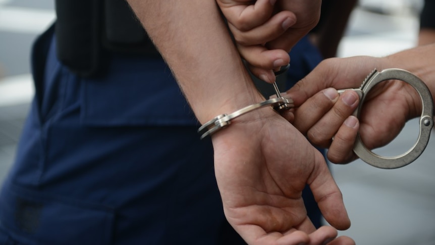 A man gets handcuffs locked on him, wearing navy pants and black top. Photo close up from side.