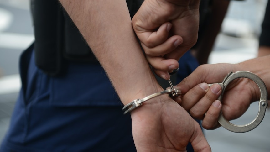 A man gets handcuffs locked on him, wearing navy pants and black top. Photo close up from side.