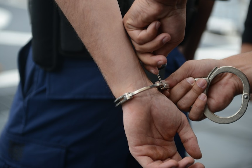 A man gets handcuffs locked on him, wearing navy pants and black top. Photo close up from side.