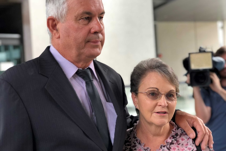 A middle-aged couple stand outside a building. The man is tall and the woman is short.