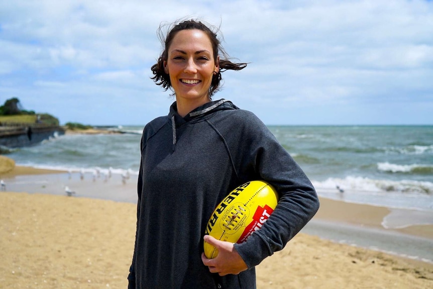 AFLW player Meg Downie stands on a beach holding a yellow Sherrin.