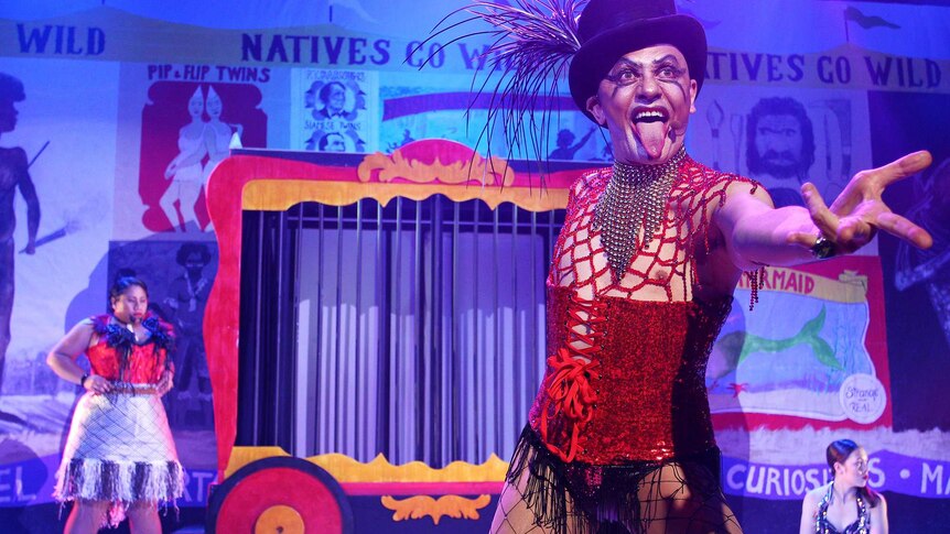 Cabaret set with performer in foreground wearing fishnets, red corset and black top hat with feathers, sticking tongue out.