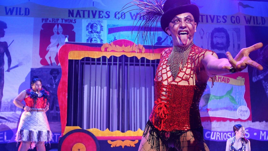 Cabaret set with performer in foreground wearing fishnets, red corset and black top hat with feathers, sticking tongue out.