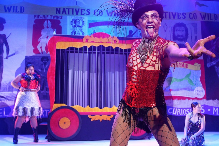 Cabaret set with performer in foreground wearing fishnets, red corset and black top hat with feathers, sticking tongue out.