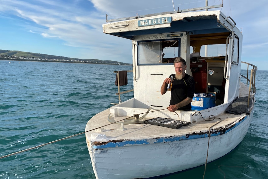 A man with a beard smiles at the camera while drinking a beer on an old wooden boat named Margrel