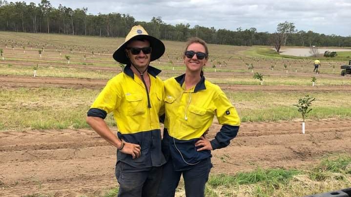 A young couple in high visibility workwear smile for a photo while working on a macadamia farm.