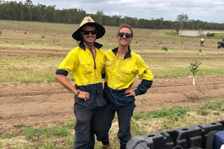 A young couple in high visibility workwear smile for a photo while working on a macadamia farm.