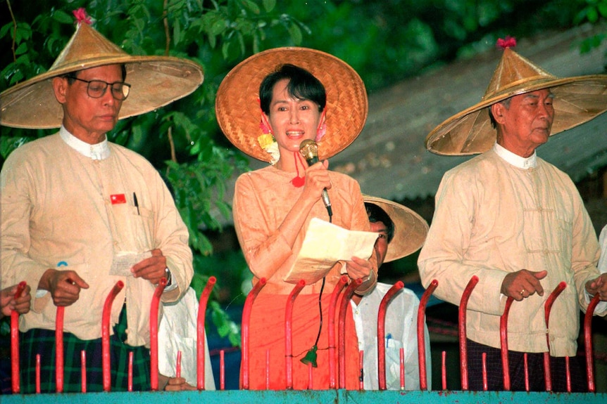 A woman with dark hair and wearing an orange dress with a straw hat speaks into a microphone with two men on either side.