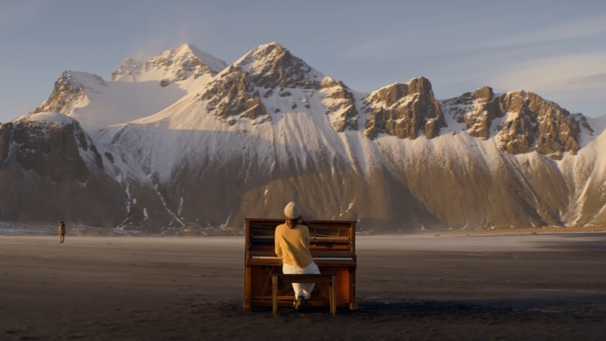 a woman sits at an upright piano in a wide open plain, tall mountains ahead of her.