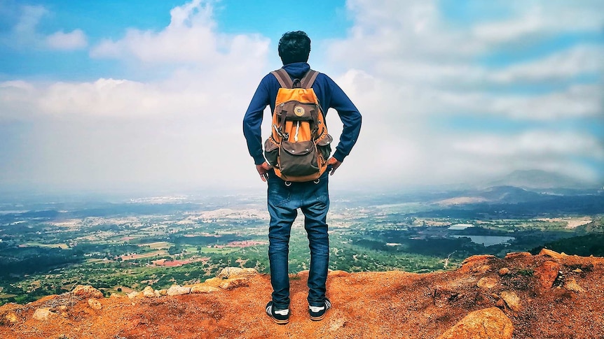Man with backpack overlooking panoramic view of mountains, greenery and buildings for a story about travelling without family.