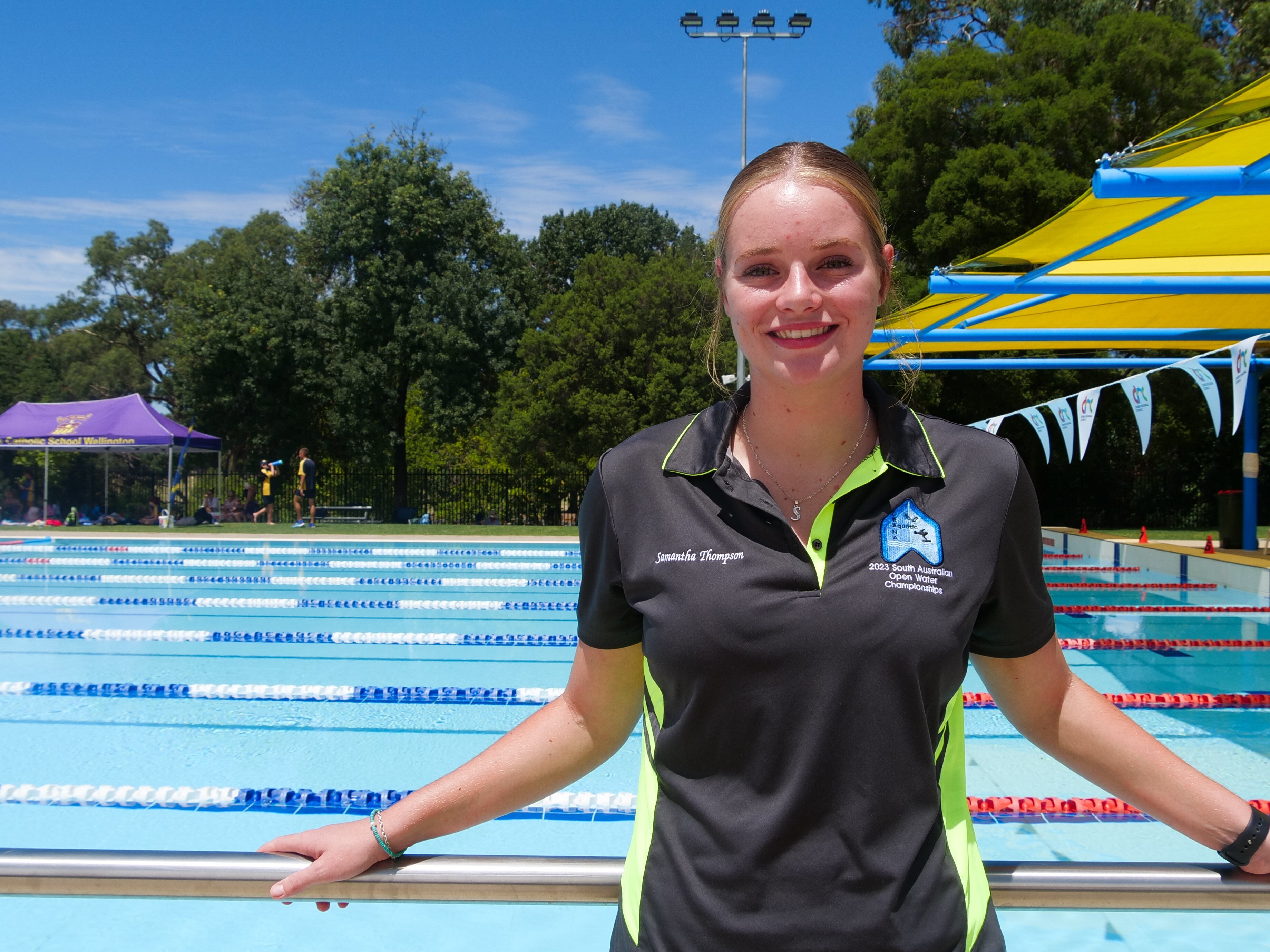 A young woman in a black and green polo shirt stands in front of an outdoor swimming pool.