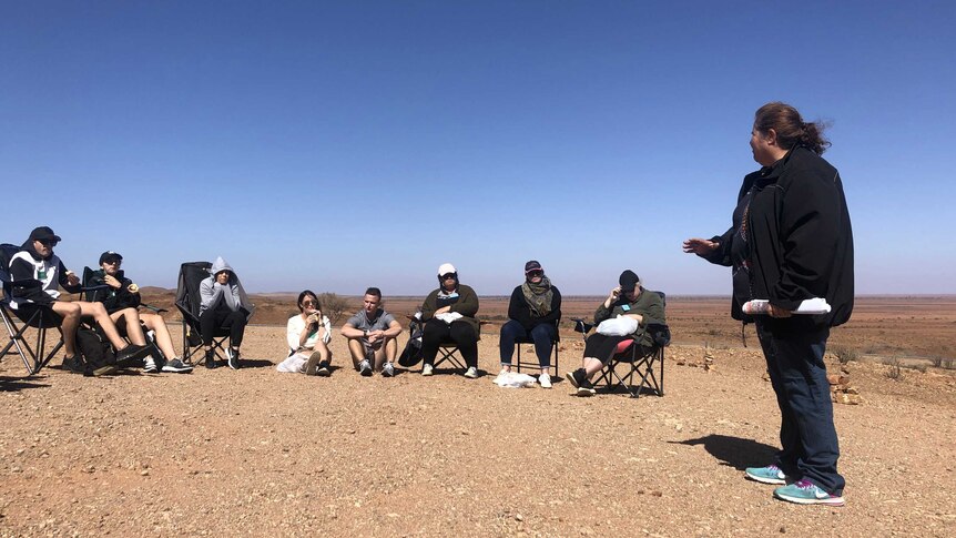 An Aboriginal woman talks to a large group on a hill overlooking a desert plain.