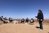 An Aboriginal woman talks to a large group on a hill overlooking a desert plain.