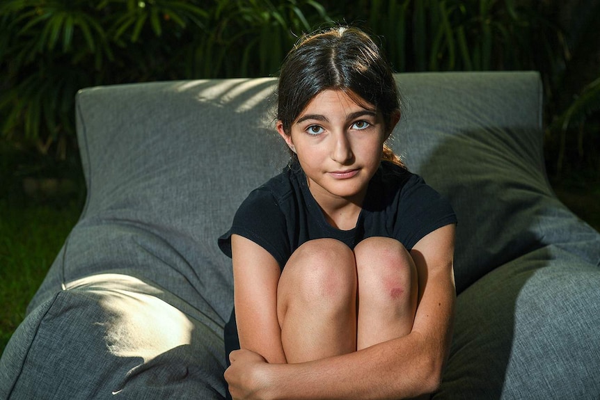 Maddy Steinmetz-Lynton aged 12, sitting on beanbag clutching her knees in her backyard looking worried, wearing a t-shirt.