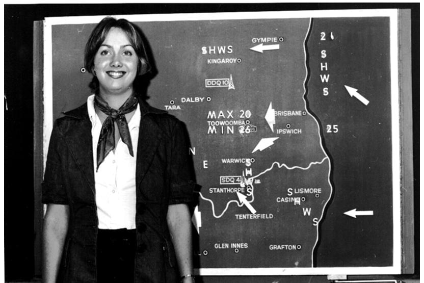Black and white photo of a woman standing in front of a map of Queensland and northern NSW with weather information stuck on.