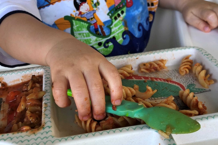 One year old baby shown from nose down with pirate bib holds a spoon on a bento plate that has pasta