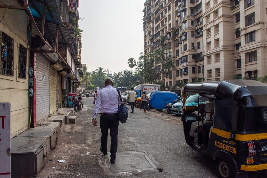 A man walking on a road next to slums with wealthier apartments in the background