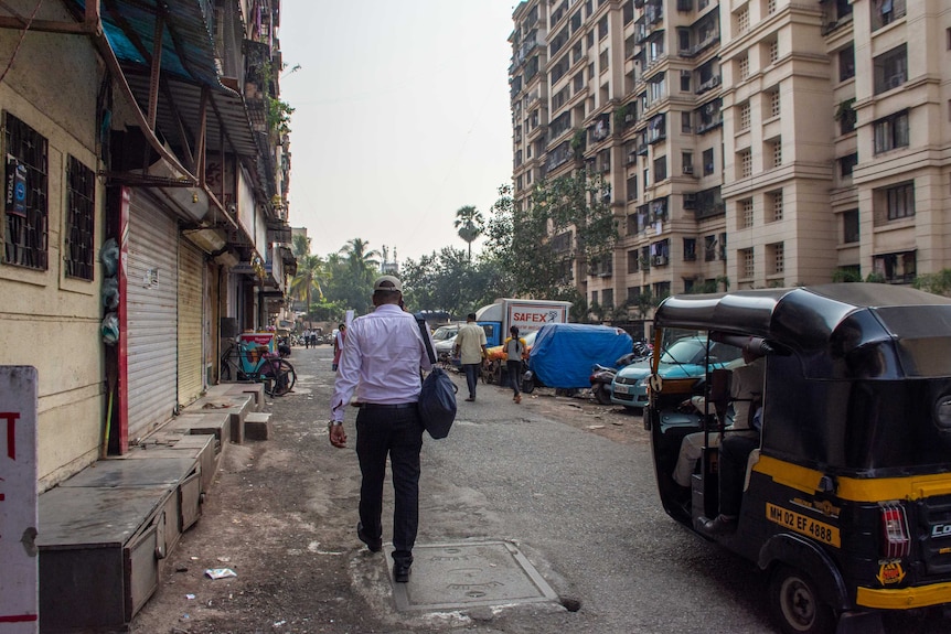A man walking on a road next to slums with wealthier apartments in the background