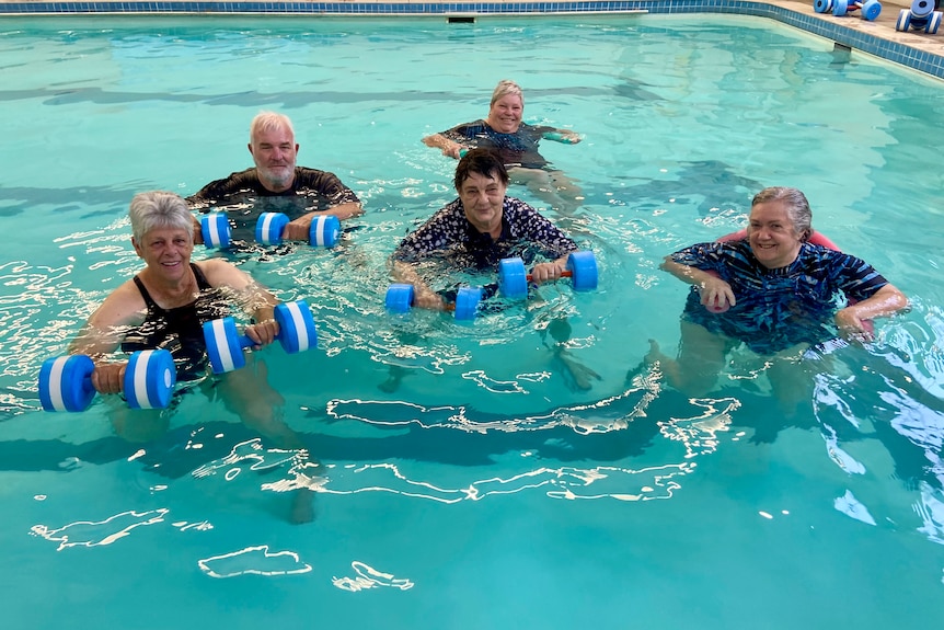 A group of elderly people smile to the camera whilst holding blue and white foam dumbells.