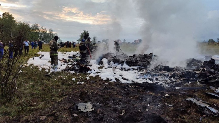 The smoking wreckage of a jet with firefighters standing around it
