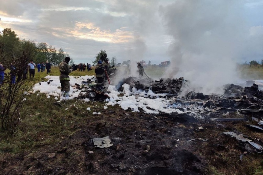 The smoking wreckage of a jet with firefighters standing around it